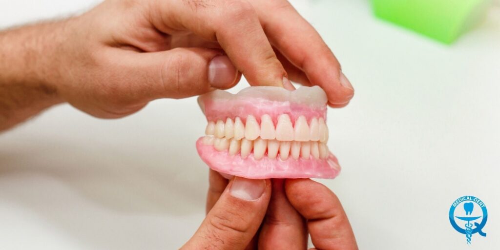 Na fotografii sú ruky držiace umelú čeľusť s radom rovných bielych zubov. Na pozadí rozmazaného svetla a lekárskych log je vidieť prsty, ktoré jemne držia model zubnej protézy. Obrázok naznačuje súvislosť s oblasťou zubného lekárstva alebo zubnej protetiky.