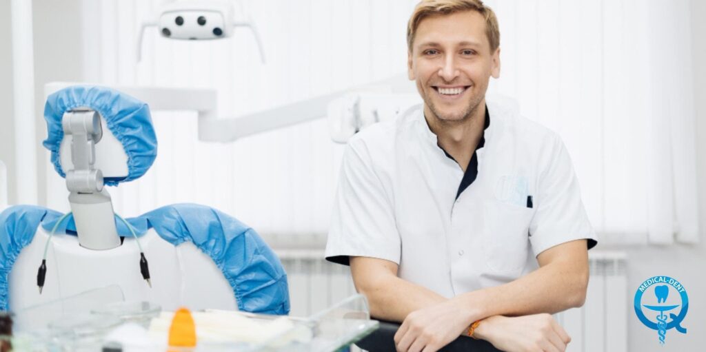 Imaginea prezintă un dentist zâmbitor, în halat medical alb, așezat la un scaun dentar modern. Echipamentul dentar profesional este vizibil, inclusiv o lampă de tratament și instrumente dentare cu huse de igienă albastre de unică folosință. Dentistul prezintă o atitudine prietenoasă și profesionistă în cadrul unui cabinet stomatologic steril.