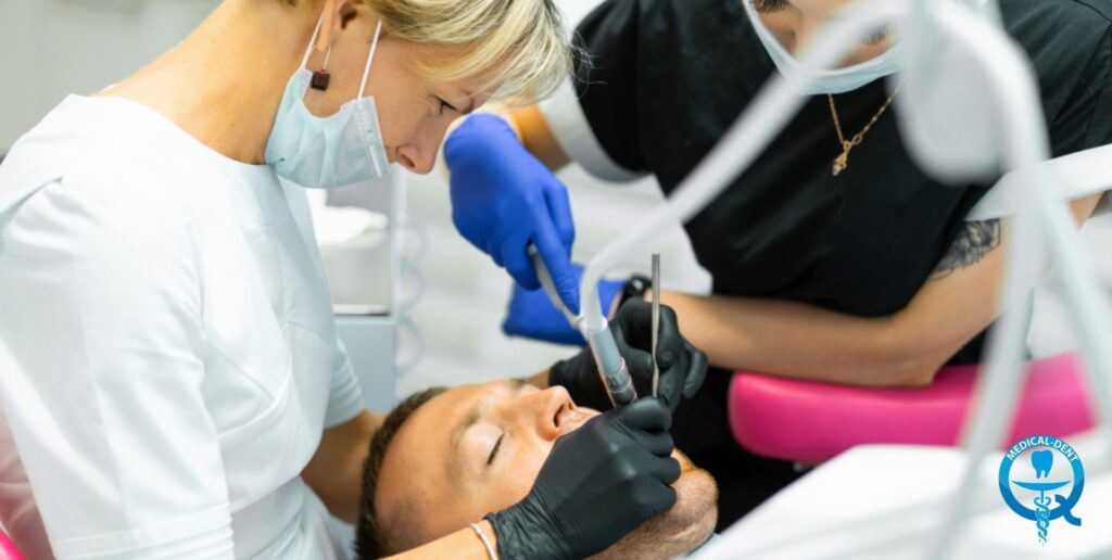 The image shows a dentist putting on blue gloves and leaning over a patient lying in a dental chair. The dentist is wearing a white protective gown and wearing headphones over his ears. The patient has his eyes closed and his head lowered while the dentist prepares to perform the dental procedure.