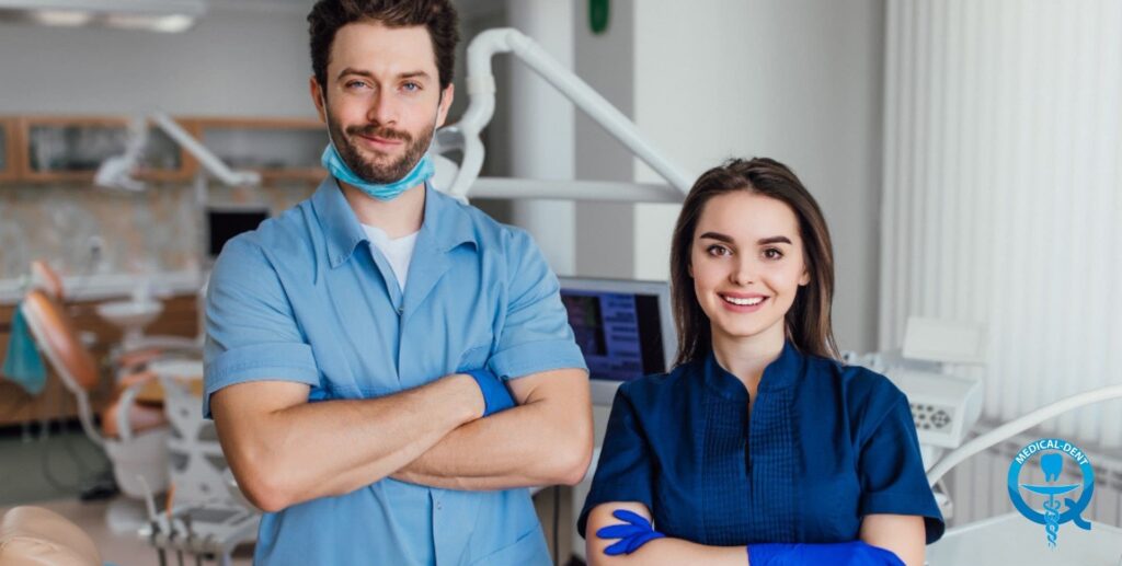 The image shows two smiling young dentists in blue uniforms standing in a professional dental office. It is equipped with a modern dental chair and medical equipment visible in the background. The male dentist is wearing a protective mask and the female is wearing blue gloves. Both have a friendly, confident demeanour, which suggests their professional competence.