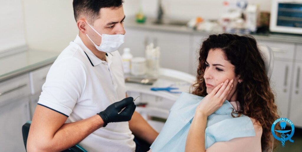 The photograph shows a dentist in a white surgical apron and mask putting on gloves in the presence of a patient sitting in a dental chair in a dental office. The woman is wearing a blue T-shirt and looks concerned, covering her mouth with her hand. The background is the interior of a typical dental office with various medical instruments.