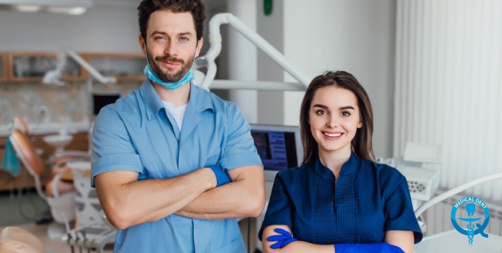 The painting depicts two smiling dentists in blue medical uniforms sitting in a dentist's office. The man has a beard and gloves on, while the woman is dressed in a jumper. Behind them, equipment and furniture typical of a dental office can be seen.