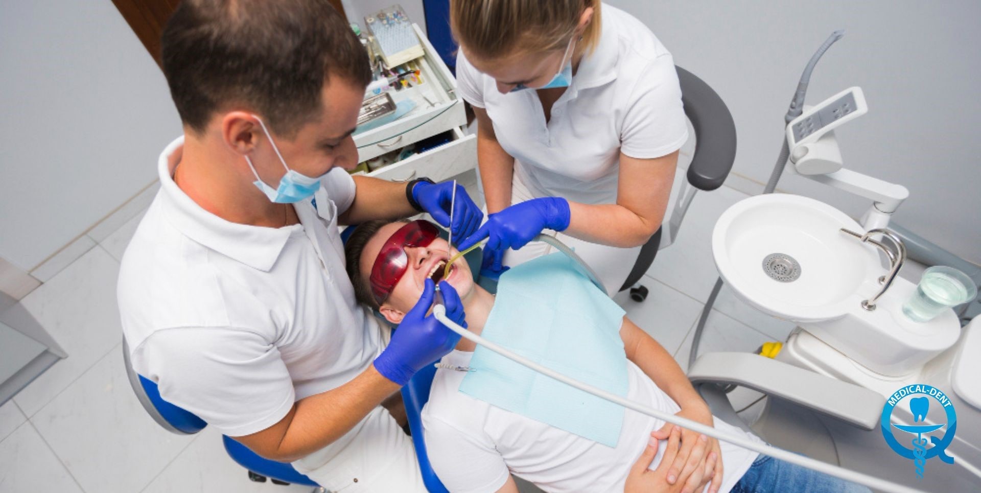 The image shows two people in medical garb performing a procedure to treat a dry socket. The patient is lying on a couch while a dentist wearing blue gloves performs the procedure in the patient's mouth. Also visible are some of the dental instruments and equipment in the treatment room.