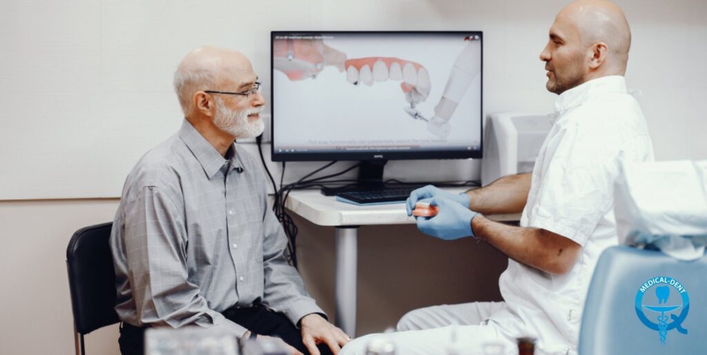 The image shows a dentist presenting a virtual denture to a patient on a large screen. The dentist, an elderly man with a grey beard, sits and watches the presentation while his colleague, a younger man in a white apron and wearing gloves, holds the instructions and explains the procedure for fitting the denture. The medical logo is visible in the foreground.