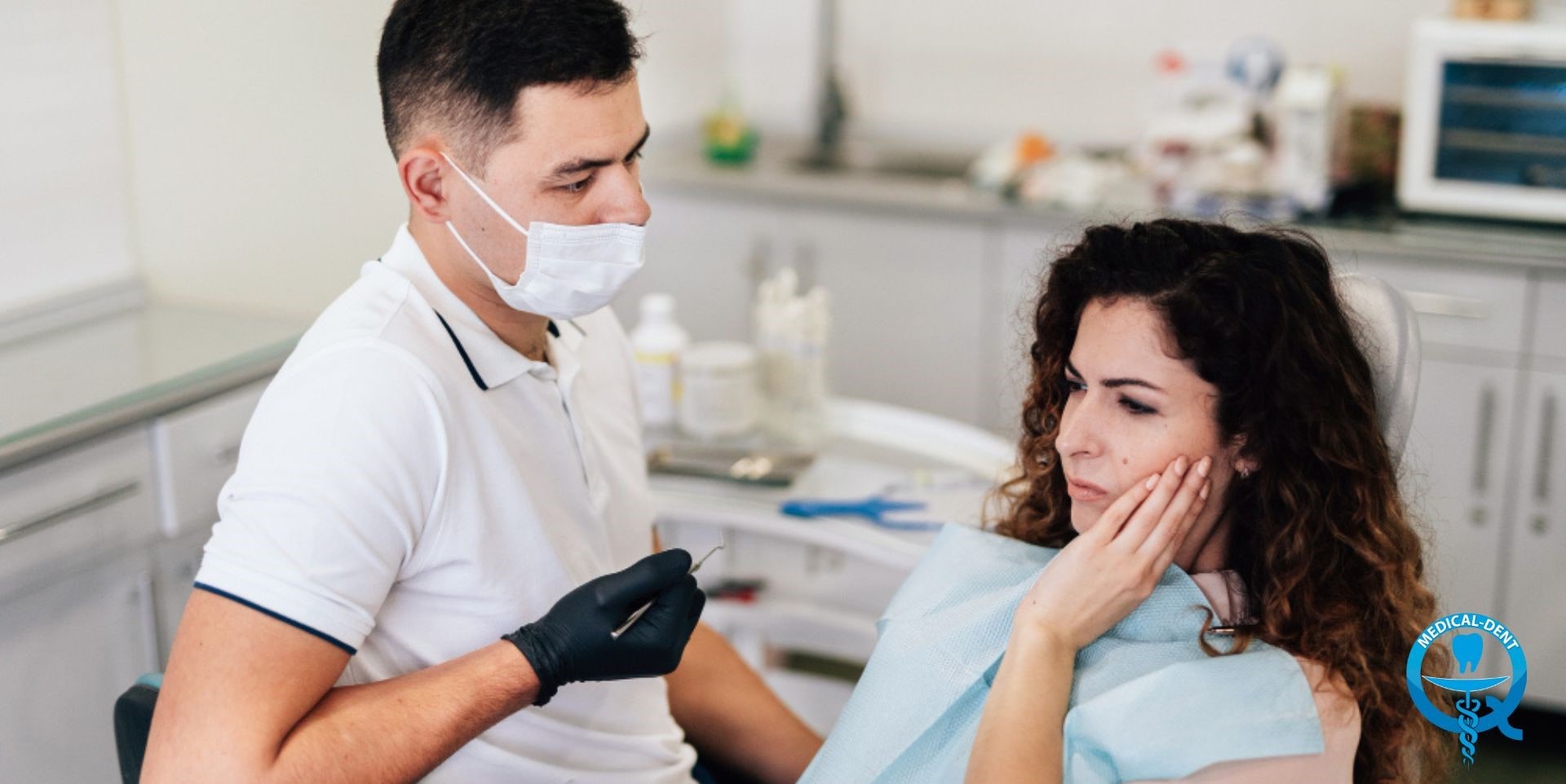 The image shows a dentist wearing a mask and gloves during a dental procedure performed on a female patient. The woman has curly hair, is holding her hand to her mouth and looks concerned. There are dental paraphernalia on the tabletop and a computer can be seen in the background. The scene takes place in a dentist's office.