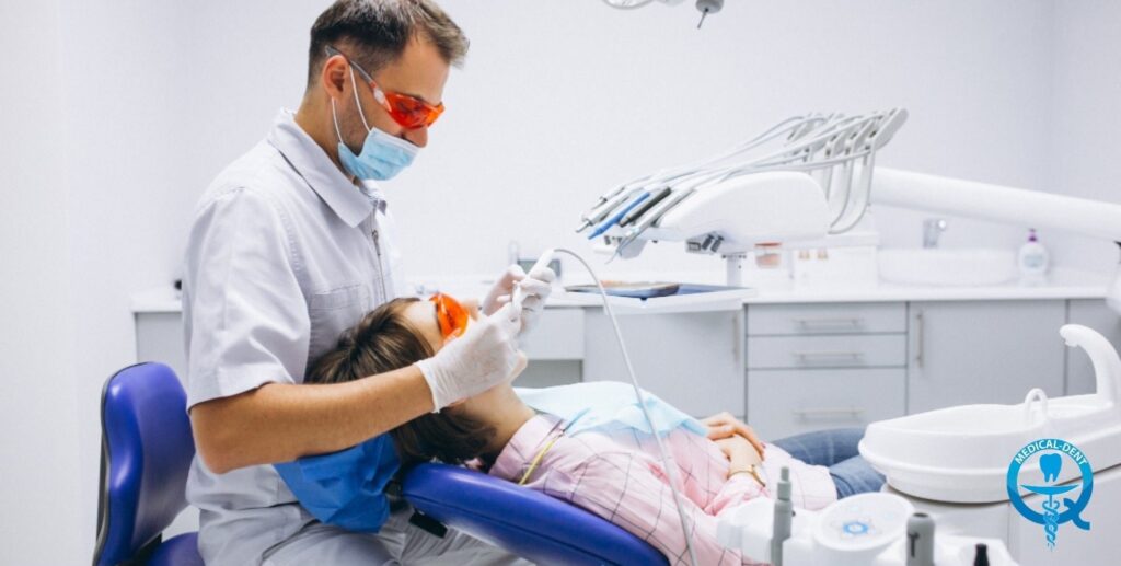 The painting shows a dentist wearing a white apron, mask and orange safety goggles performing a dental procedure on a patient lying in a dental chair. On the table beside her are dental instruments. The identities of the people in the picture have not been revealed.