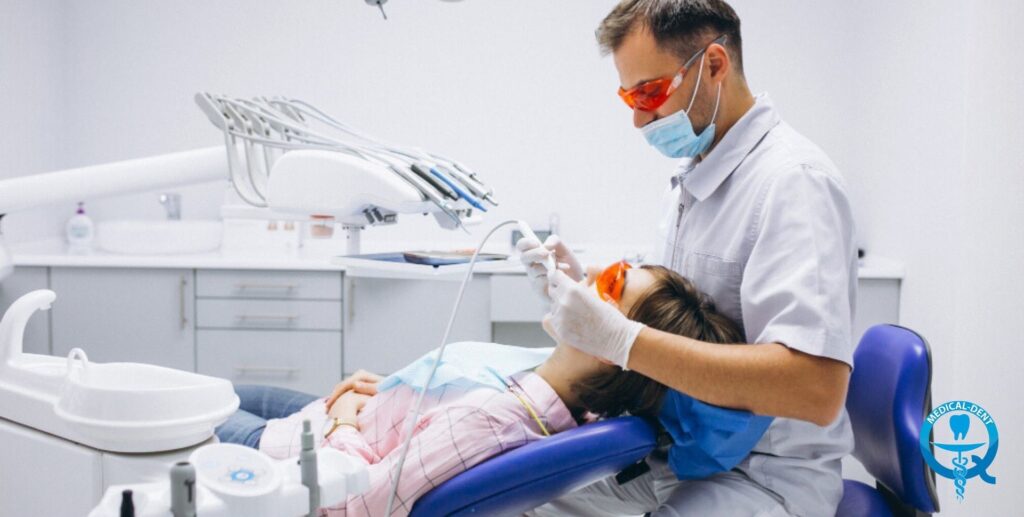 Fotografia înfățișează un medic dentist cu protecție completă - mască, ochelari de protecție și șorț - efectuând o procedură dentară pe un pacient întins pe un scaun dentar. Echipamentul profesional și instrumentele dentare aranjate în jurul scaunului pot fi văzute.