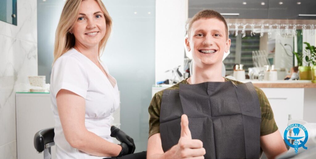 The photograph shows a smiling blonde woman and a man in a dental or orthodontic practice. Both appear to be employees of the clinic and are posing in a friendly manner. The equipment of the practice can be seen, including a treatment chair and a bright kitchen in the background.