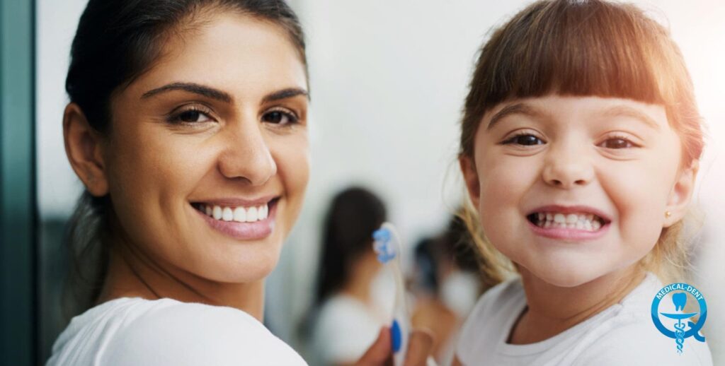 The photo shows a smiling dentist and a smiling little girl, presumably her patient. The dentist is wearing a white medical apron and the child is wearing a white T-shirt. They are sitting next to each other and look happy after a dental visit.