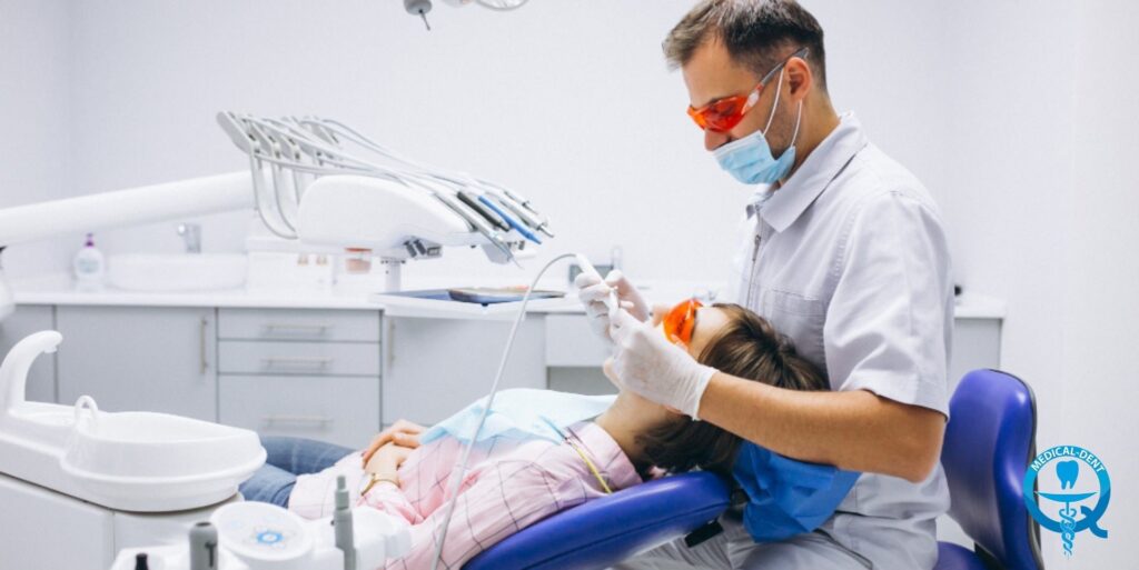 Imaginea prezintă un dentist purtând mască și ochelari de protecție care efectuează o procedură dentară pe un pacient întins pe un scaun dentar. Instrumentele dentistului și echipamentul cabinetului dentar sunt vizibile.