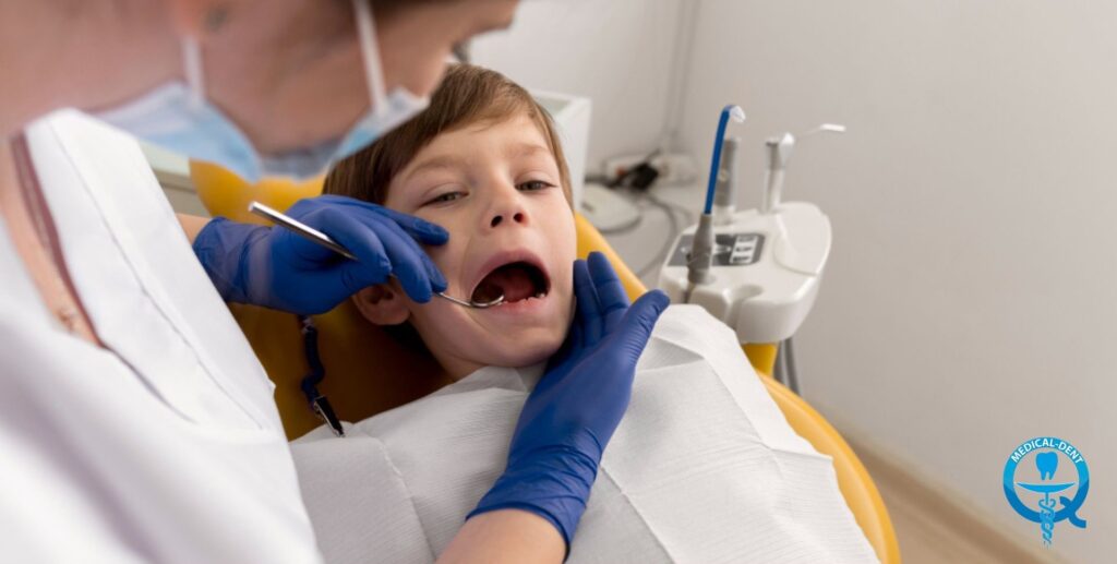 The photo shows a young child undergoing a dental procedure. The child can be seen croaking with his mouth open and a sleepy face as the blue-gloved dentist performs the procedure. The dental instruments can be seen in the foreground, and the dental munit in the background.