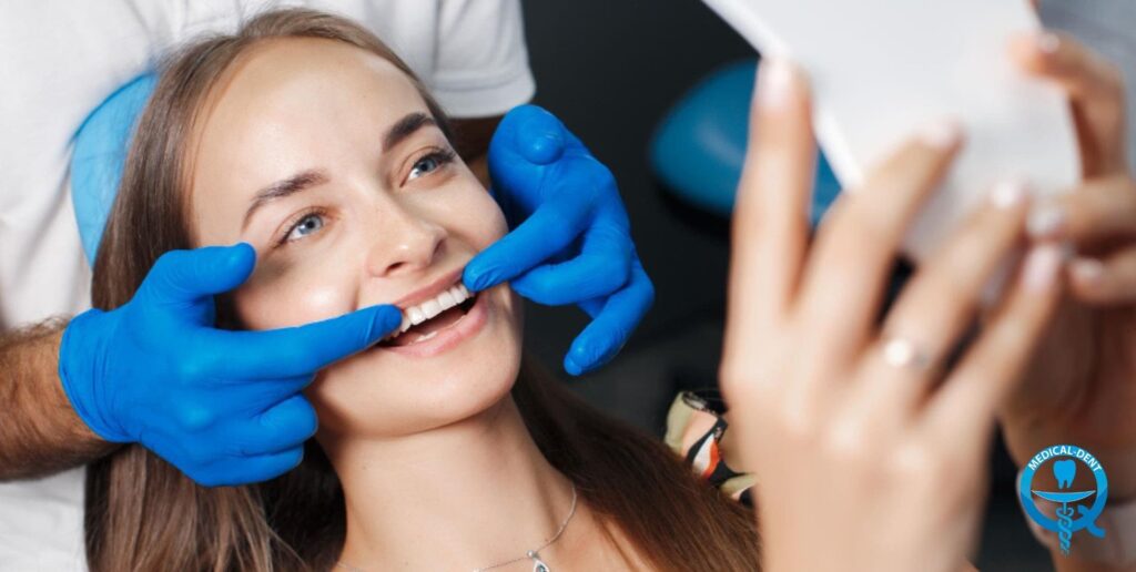 The photograph shows a young woman during a visit to the dentist. She is dressed in a white blouse and the dentist, wearing blue gloves, is examining her teeth by gently pushing back her lower lip. She is smiling, but her face is not visible up close to maintain privacy. The dental logo on the photo is also visible.