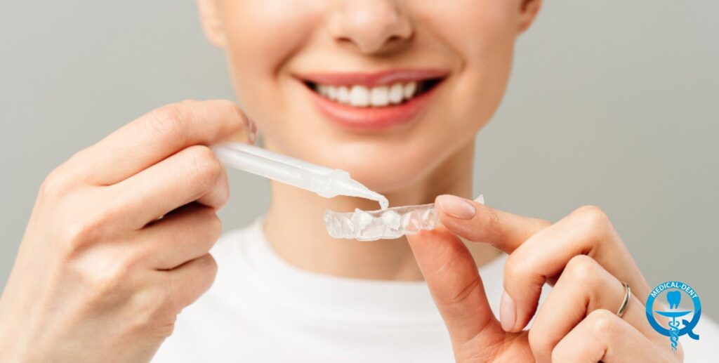 The image shows a smiling person holding a plastic teeth whitening tray on the lower teeth. The person has white teeth and is holding his hands in front of his mouth to put the whitening tray on. The image promotes a home teeth whitening kit.