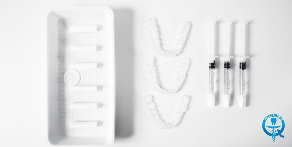 The photograph shows a kit for home teeth whitening. Visible are a white container, presumably with a whitening gel or other preparation, a flexible tube and three narrow cylinders with a thick white substance, presumably gel applicators for the teeth. In the lower right corner is the medical logo.